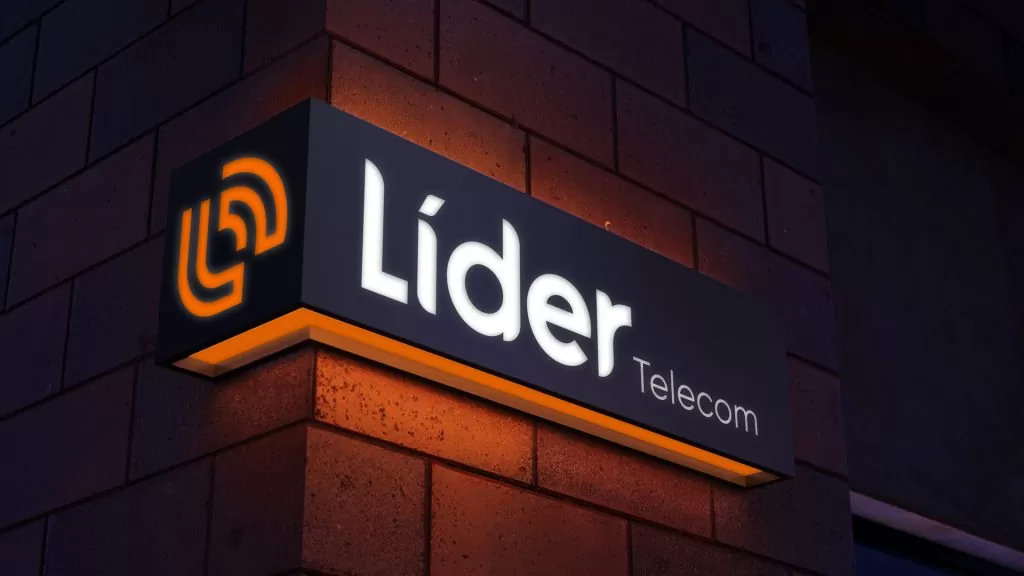 Painel luminoso de fachada, à noite, com o símbolo laranja, a palavra Líder em branco e a palavra Telecom, sobre parede de tijolos com faixa de luz laranja